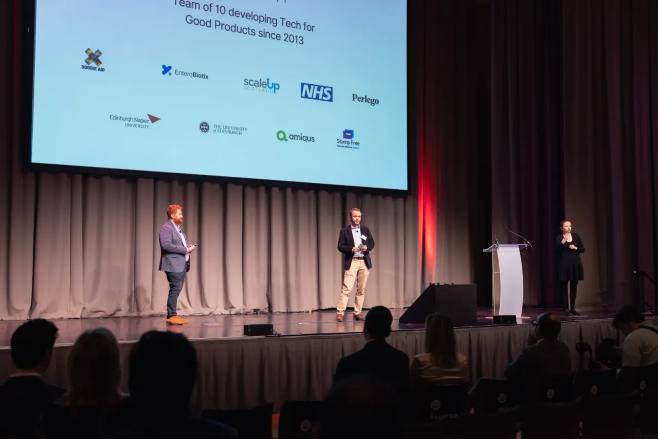 Two of the team presenting on stage to an audience, with a sign language interpreter.